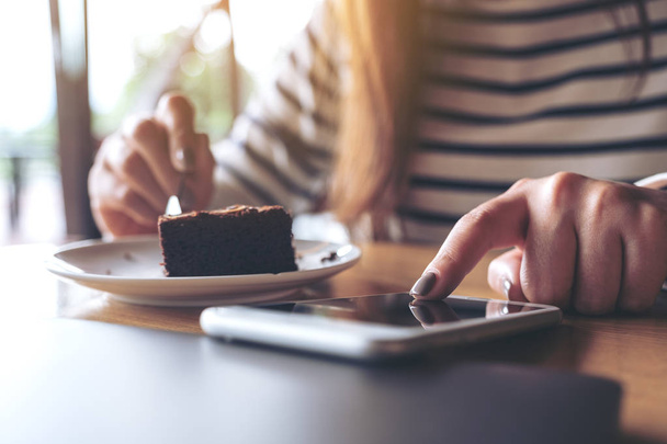 Closeup image of a woman using and touching a smart phone while eating brownie cake in cafe - Фото, изображение
