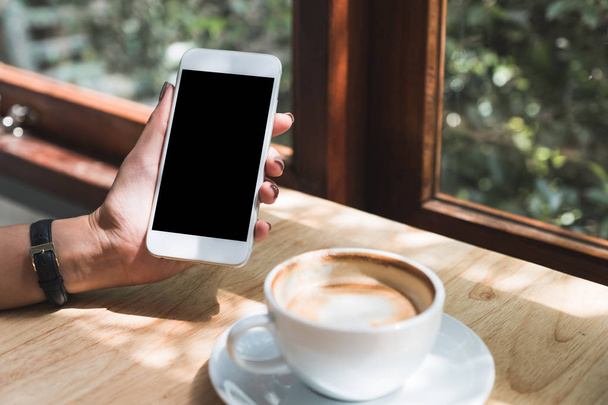 Mockup image of a hand holding and showing white mobile phone with blank black desktop screen with coffee cup on wooden table in cafe - Foto, afbeelding