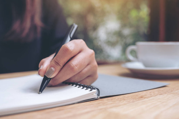 Szoros kép egy nő kézírásáról egy fehér üres noteszre, kávéscsészével a fa asztalon.  - Fotó, kép