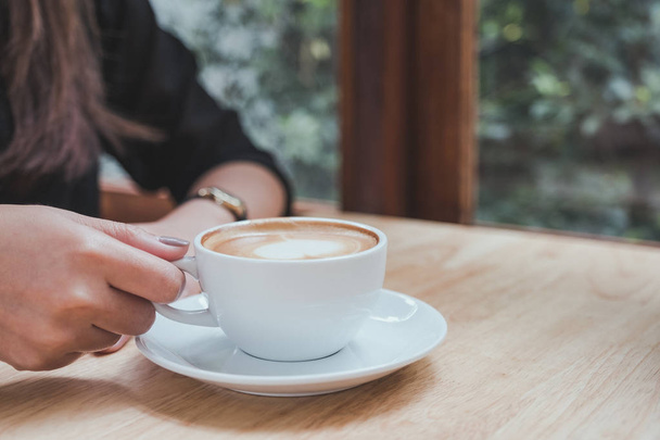 Nahaufnahme einer Frau mit einer weißen Tasse heißen Kaffees auf einem Holztisch im Café - Foto, Bild