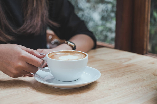 Крупный план женщины, держащей чашку белого горячего кофе на деревянном столе в кафе - Фото, изображение