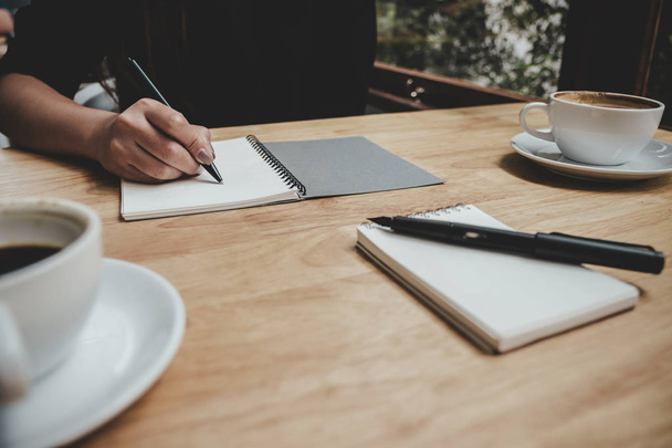 Imagen de cerca de la mano de una mujer escribiendo en un cuaderno blanco en blanco con taza de café en la mesa de madera
  - Foto, Imagen