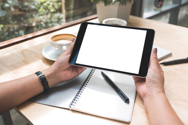 Imagem Mockup de mãos de uma mulher segurando pc tablet preto com tela branca em branco com notebook e xícara de café na mesa
  - Foto, Imagem