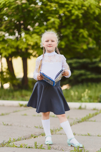 little blonde girl in full length with pigtails reading a book outdoors - Фото, изображение