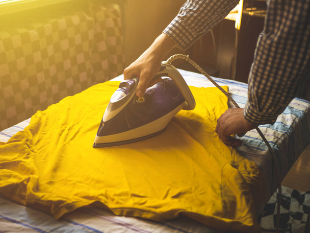 Close-up van iemands handen Strijkservice t-shirt in de Zomerochtend in slaapkamer met gele zon - Foto, afbeelding