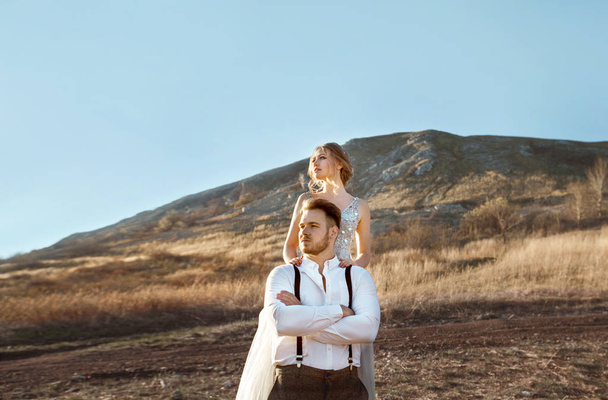 beautiful young couple man and woman standing against the background of a mountain and blue sky in wedding dresses in summer at sunset - 写真・画像