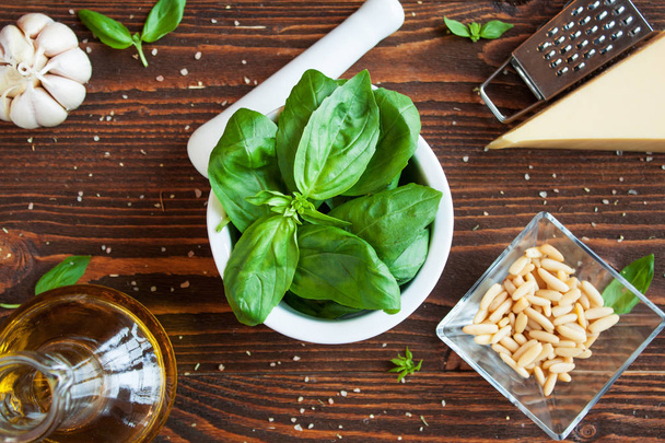 Basil leaves in mortar, olive oil, pine nuts, garlic, parmesan cheese on rustic wooden table. Ingredients for basil pesto preparation - Photo, Image