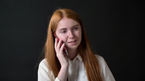 Young charming woman with long ginger hair talking on cell phone, girl in white shirt isolated on black background - Video, Çekim