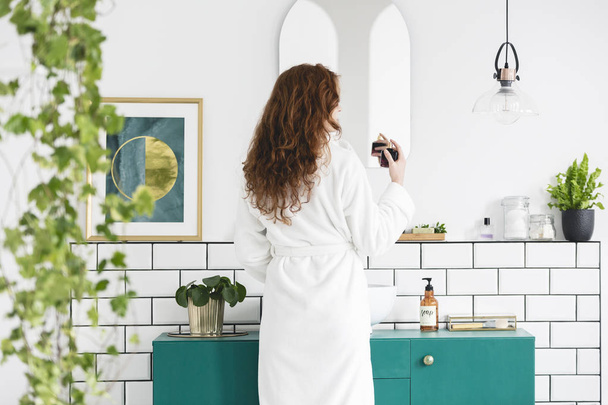 Woman in front of mirror in white bathroom interior with poster, plants and green cabinet. Real photo - Foto, Imagen