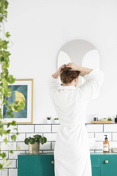 Mujer en bata blanca delante del espejo en el interior del baño con planta y póster. Foto real
 - Foto, Imagen