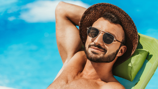 hombre guapo en gafas de sol y sombrero de paja tomar el sol en la cama al lado de la piscina
 - Foto, Imagen