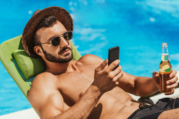 man in sunglasses using smartphone and holding bottle of beer on sunbed at poolside - Fotoğraf, Görsel