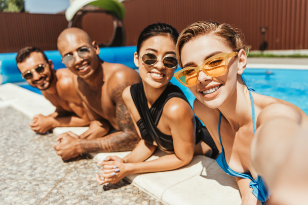 молоді усміхнені мультикультурні люди в купальниках і сонцезахисних окулярах позують у басейні
 - Фото, зображення