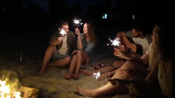 Φίλοι στην παραλία με βεγγαλικά γιορτάζει το σούρουπο ηλιοβασίλεμα. Νέοι άνδρες και γυναίκες κάθονται γύρω από την φωτιά με Βεγγάλη φώτα και τραγουδούν. Γενειοφόρος hipster παίζει κιθάρα. - Πλάνα, βίντεο