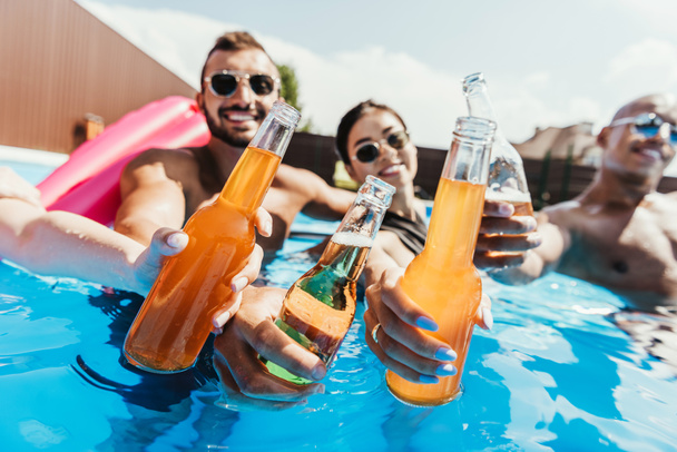 multiethnic friends clinking with bottles on beer in swimming pool - Fotografie, Obrázek