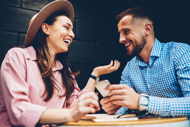 Cheerful male and female friends enjoying free time together joking and share multimedia in networks during meeting in cafe, romantic couple laughing viewing funny photos on mobile phone during date - Фото, изображение