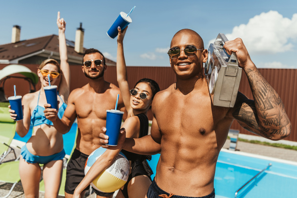 πολυεθνική τους φίλους χαμογελώντας με ποτά και ρετρό boombox δίπλα στην πισίνα - Φωτογραφία, εικόνα