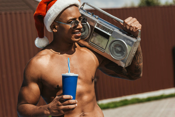 glimlachen van Afro-Amerikaanse man in kerstmuts met drankje en retro boombox - Foto, afbeelding