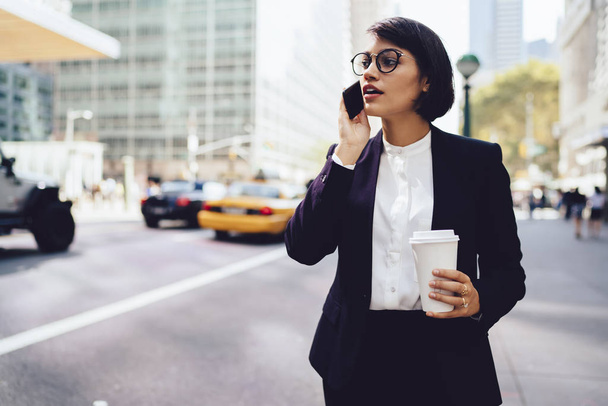 Successful financial manager in elegant wear walking in metropolis and talking on smartphone nea publicity area.Businesswoman having phone conversation about work holding coffee to go on street - Fotó, kép