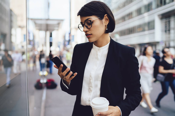 Busy owner of financial company reading income message on cellular walking on street with crowd of people.Serious female entrepreneur in elegant wear checking email on phone strolling with coffee - Photo, Image
