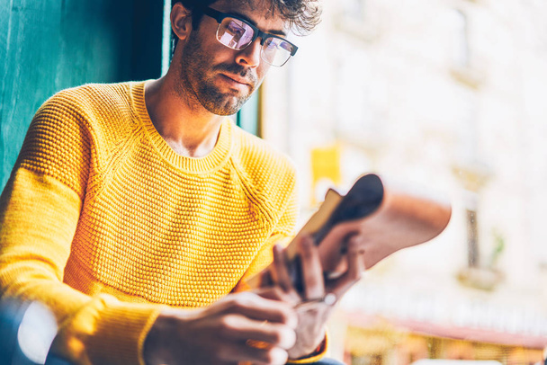 Cropped image of smart young man in optical eyegasses reading information from notepad.Pensive hipster guy dressed in yellow sweater learning text from notebook sitting in coworking space - Photo, Image