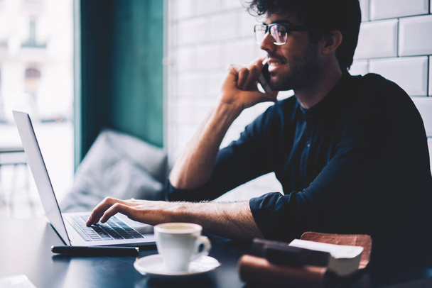 Side view of cheerful male entrepreneur in eyeglasses typing on keyboard of modern laptop device in searching information while talking on smartphone with friend sitting in coffee shop - Foto, imagen