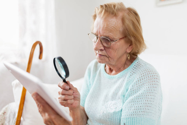 Elderly woman with glasses and loupe using a modern digital tablet - Foto, afbeelding