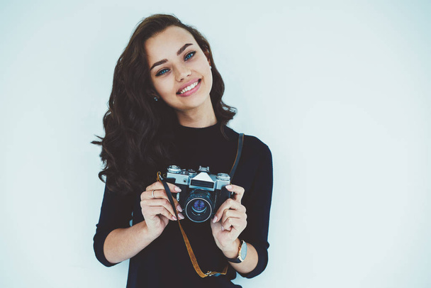 Half length portrait of smiling beautiful female working photographer and standing on white promotional background.Attractive cute young woman holding vintage camera in hand and posing on camera - Zdjęcie, obraz