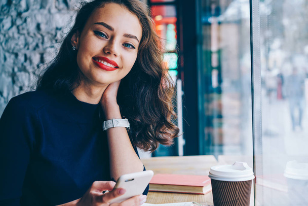Closeup portrait of attractive smiling hipster girl with brunette hair holding digital smartphone in hand and looking at camera while sitting in coffee shop.Copy space area for advertising - Photo, image