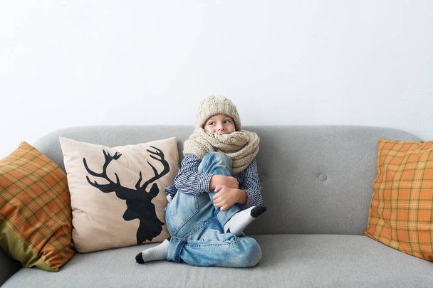 The child in a knitted cap and with a warm scarf sitting on a sofa. - Fotoğraf, Görsel