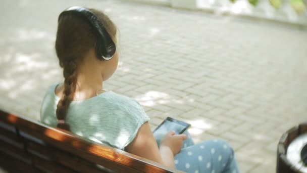 Little fat girl with a tablet PC and headphones sitting on a bench listening to music or watching a video in a summer park - Felvétel, videó
