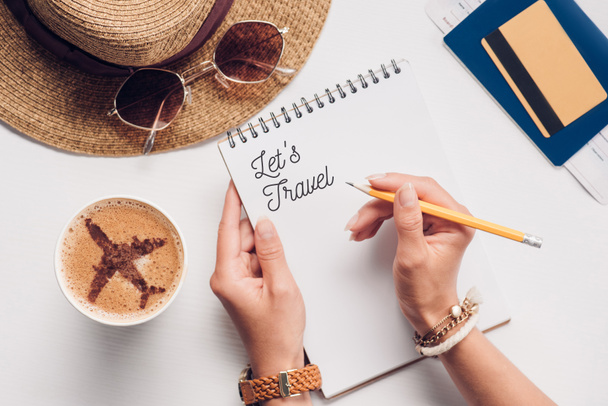 partial view of woman with notebook with lets travel lettering at tabletop with cup of coffee, straw hat, passport and ticket, vacation concept - Photo, Image