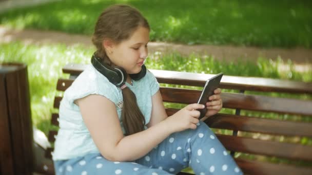 Little fat girl with a tablet PC and headphones sitting on a bench listening to music or watching a video in a summer park - Πλάνα, βίντεο