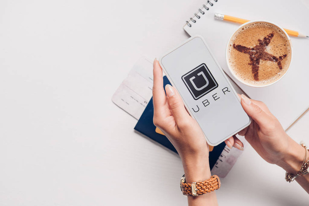 cropped shot of woman with smartphone with uber logo on screen at tabletop with cup of coffee, ticket and passport - Foto, afbeelding