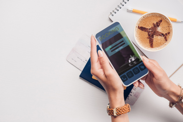 tiro recortado de mulher com smartphone com site de reserva na tela na mesa com xícara de café, bilhete e passaporte
 - Foto, Imagem