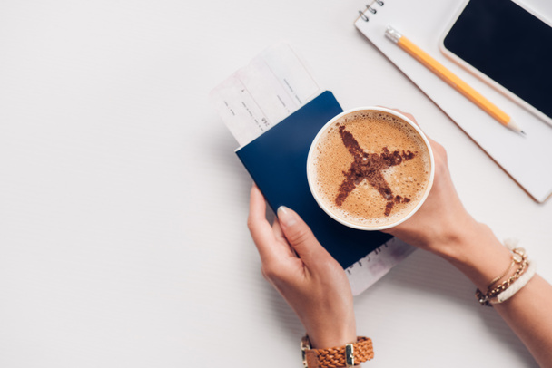 περικοπεί shot γυναίκα κρατώντας φλιτζάνι καφέ με αεροπλάνο σημάδι στο tabletop με διαβατήριο, smartphone και εισιτήριο, ταξιδεύουν έννοια - Φωτογραφία, εικόνα