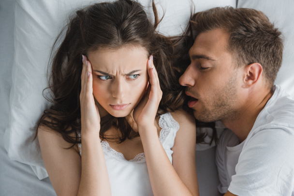 von oben betrachtet junge Frau mit Kopfschmerzen beim Anblick ihres Mannes, der im Bett schnarcht - Foto, Bild