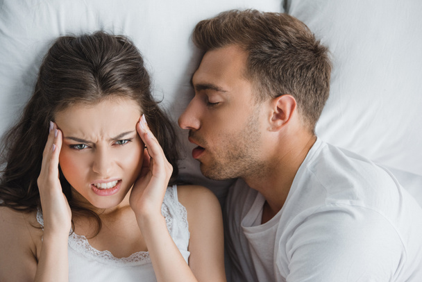 mladá naštvaná žena s bolesti hlavy při ležení chrápajících manžela v posteli  - Fotografie, Obrázek