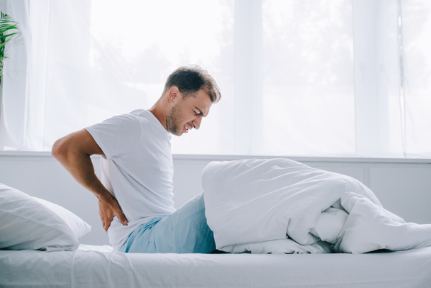 side view of young man in pajamas sitting on bed and suffering from back pain - Photo, Image