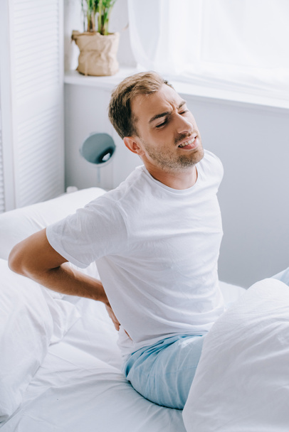 young man in pajamas sitting on bed and suffering from back pain - Photo, image