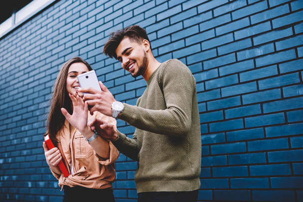 Cheerful best friends viewing funny photos in social networks via smartphone resting together outdoors, happy male and female hipsters watching videos online on mobile standing near publicity area - Fotoğraf, Görsel