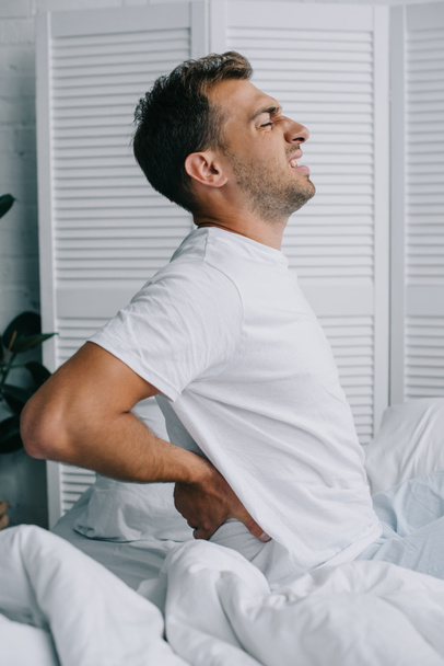 side view of young man in pajamas suffering from back pain while sitting on bed    - Photo, Image