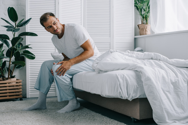 jeune homme en pyjama assis sur le lit et regardant loin tout en souffrant de douleurs au genou à la maison
 - Photo, image