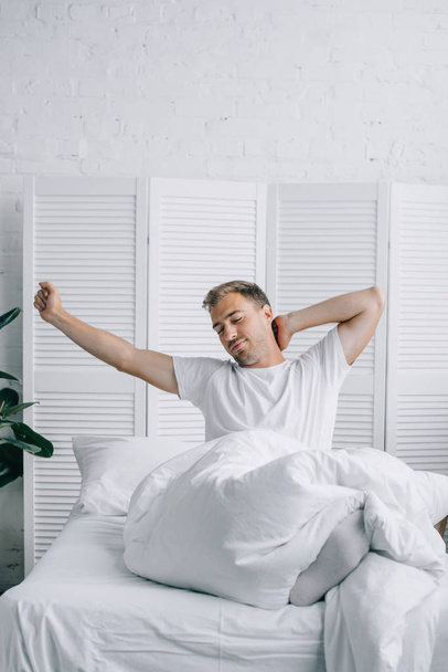 schöner junger Mann sitzt auf dem Bett und dehnt sich am Morgen - Foto, Bild