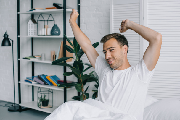 smiling young man stretching and looking away while sitting on bed - Φωτογραφία, εικόνα