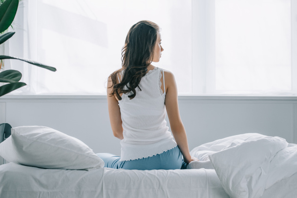 zadní pohled na mladou ženu v pyžamu, sedí na posteli a hledat dál  - Fotografie, Obrázek