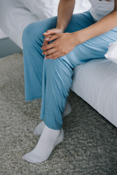 tiro cortado de mulher de pijama sentado na cama e sofrendo de dor no joelho
  - Foto, Imagem