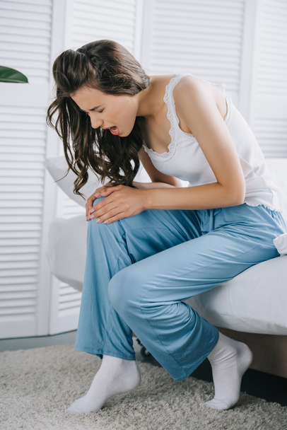 νεαρή γυναίκα στο πιτζάμες που υποφέρουν από πόνο στο γόνατο, ενώ κάθεται στο κρεβάτι στο σπίτι - Φωτογραφία, εικόνα