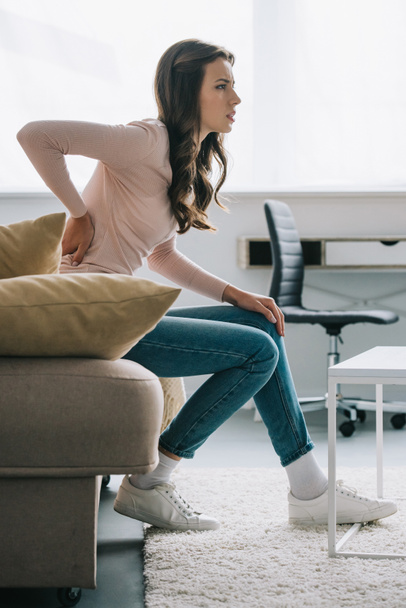 side view of young woman sitting on sofa and suffering from back pain  - Fotoğraf, Görsel
