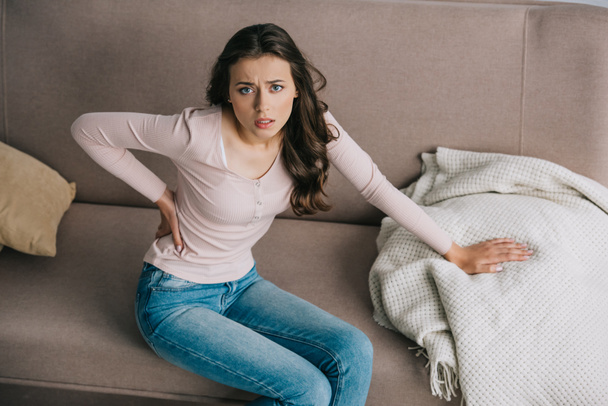 high angle view of young woman with back pain sitting on couch and looking at camera - Foto, Imagem
